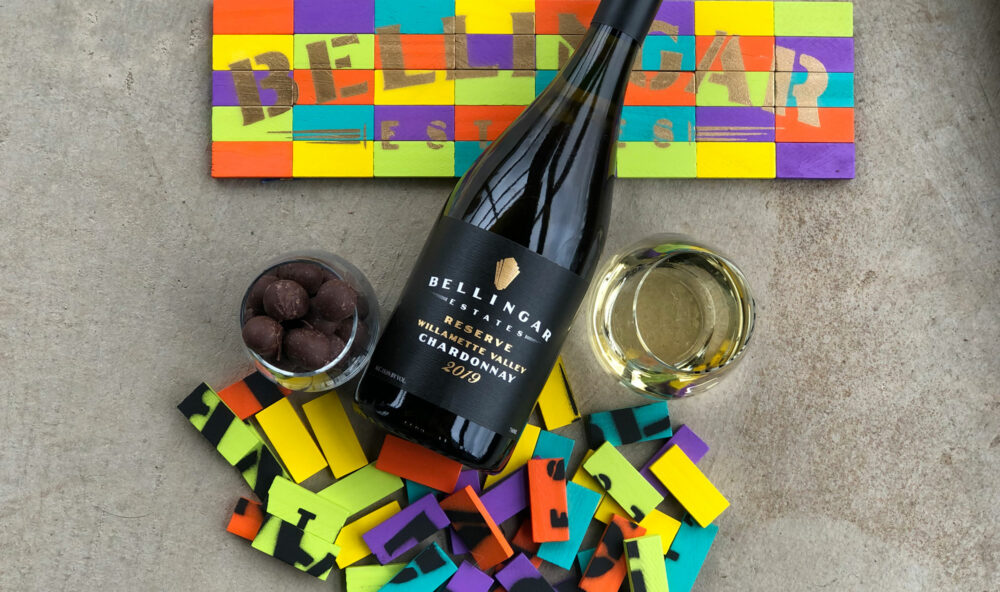 Bottle of Bellingar Chardonnay lying on its side between two glasses of wine, on top of a multicolored logo sign